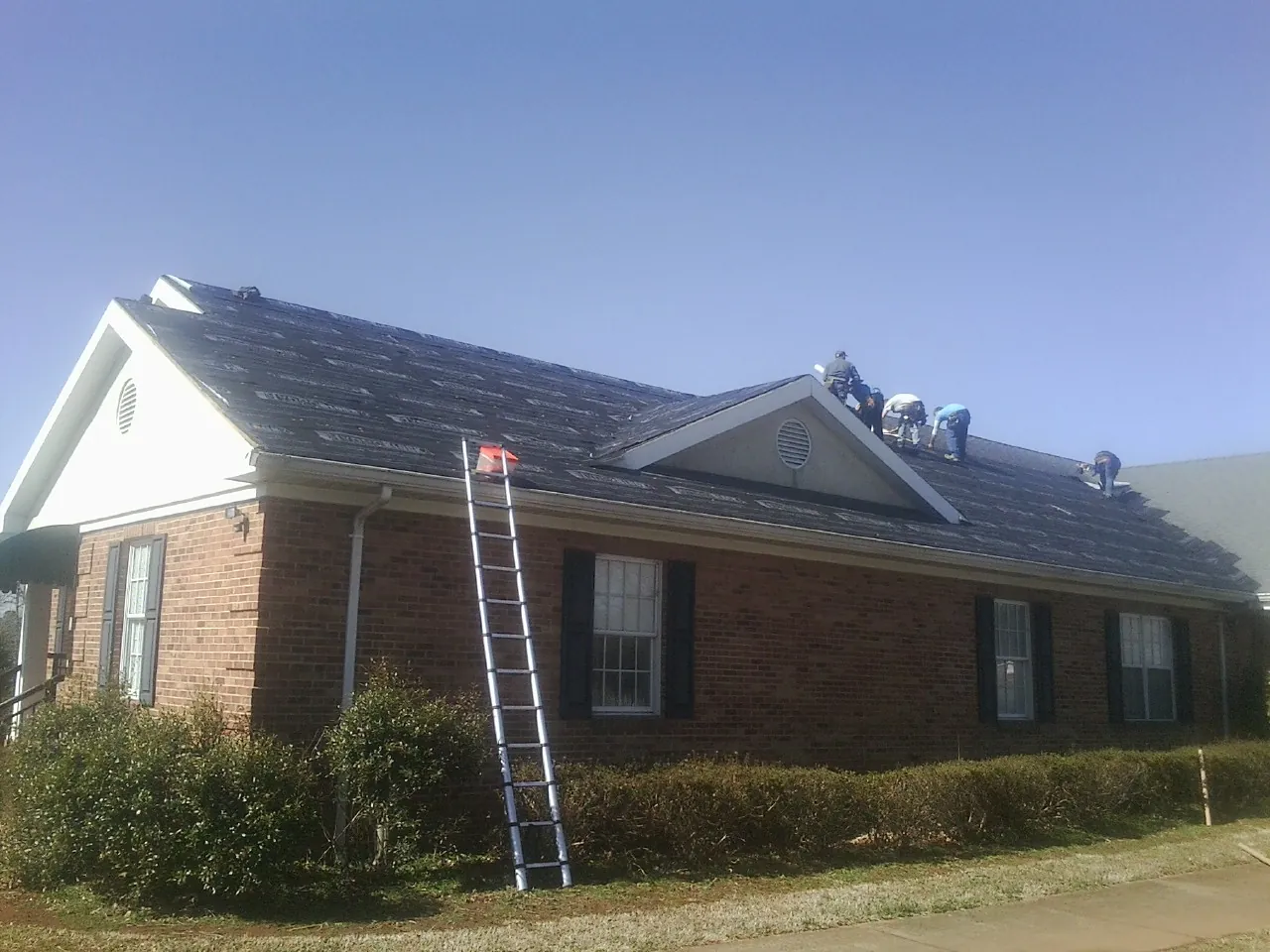 Crew tearing shingles off a brick ranch roof with a ladder set — Emergency Roof Repair work by our crew in La Prairie