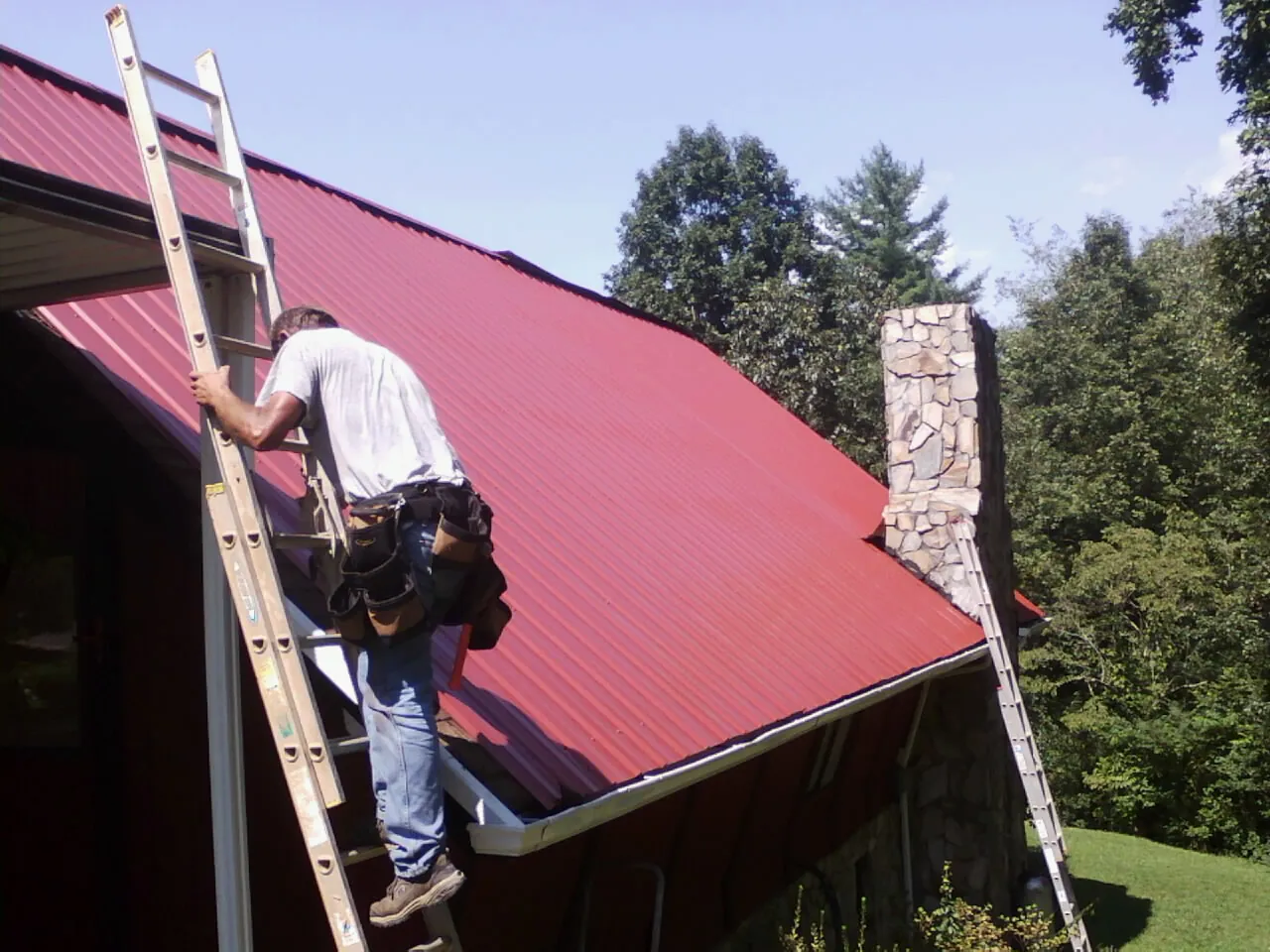 Roofer on a ladder beside a red metal roof and stone chimney — Soffit & Fascia Repair by Roofing Brothers in La Prairie